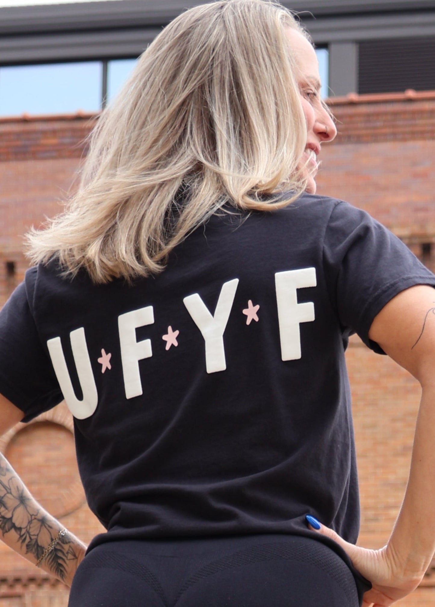 Person wearing a black t-shirt with 'U.F.Y.' text, black leggings, and white sneakers standing in front of a brick building.
