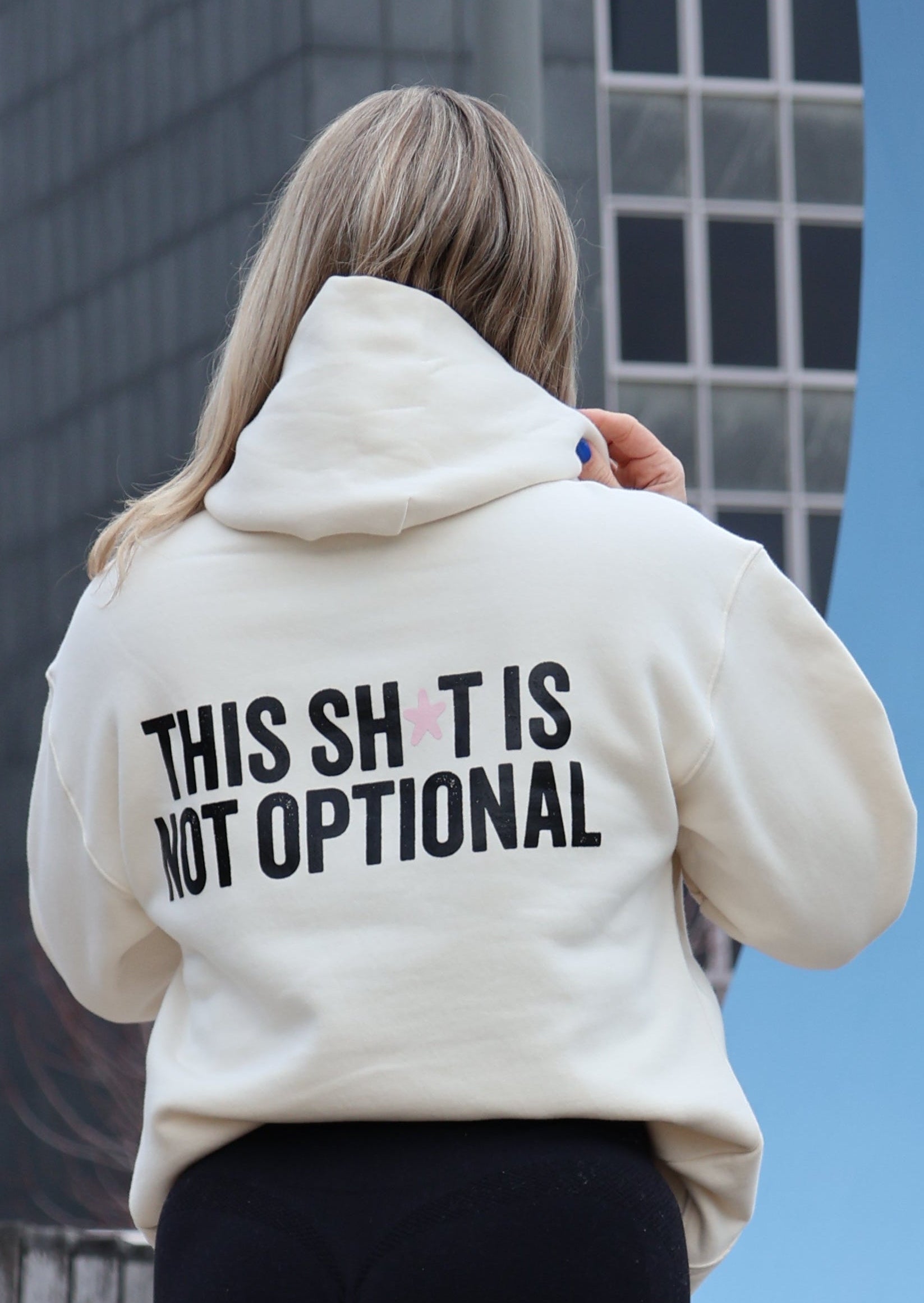 Person wearing a beige hoodie with text, standing on a sidewalk with buildings in the background