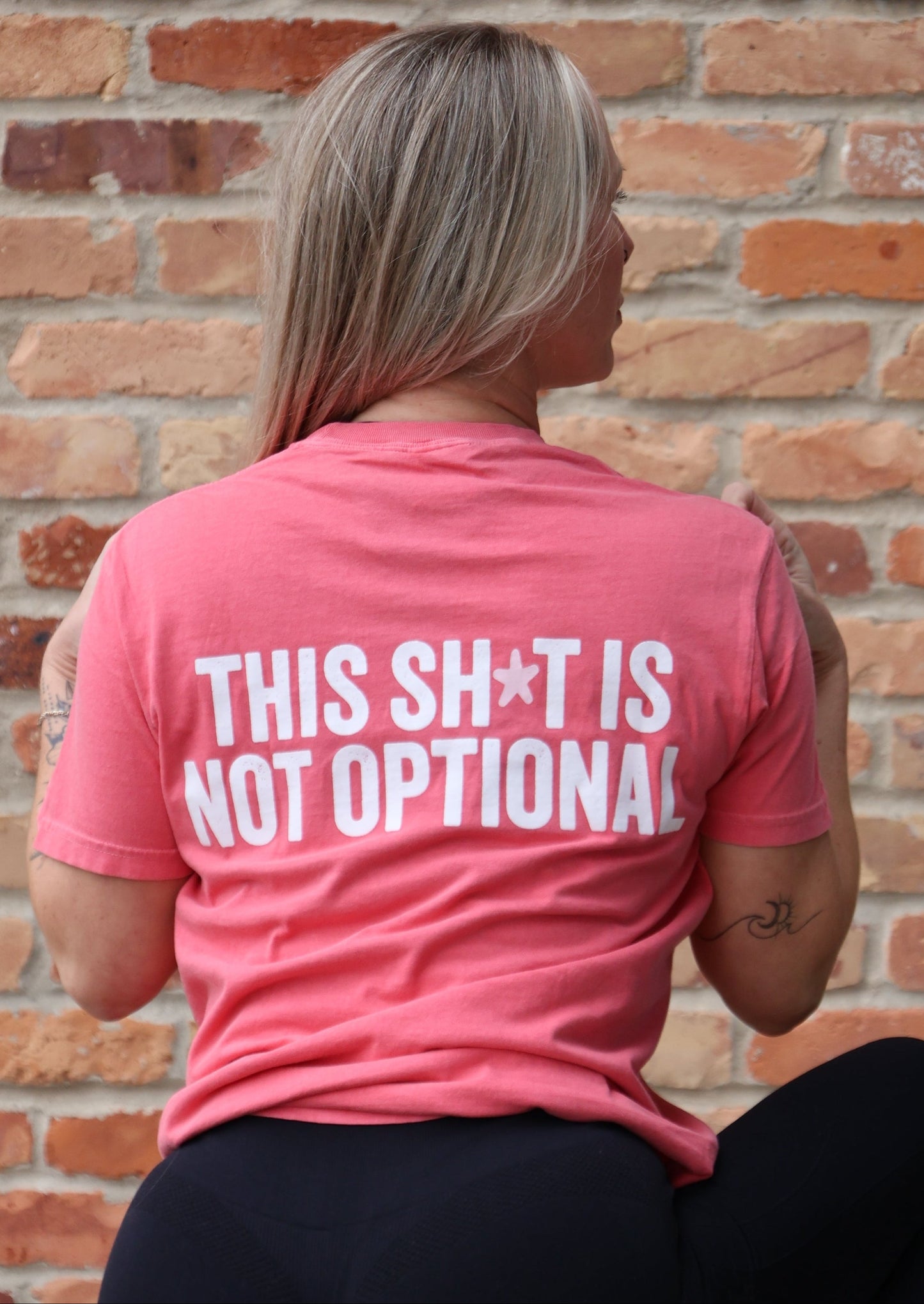 Person wearing a pink shirt with text, standing in front of a brick wall.