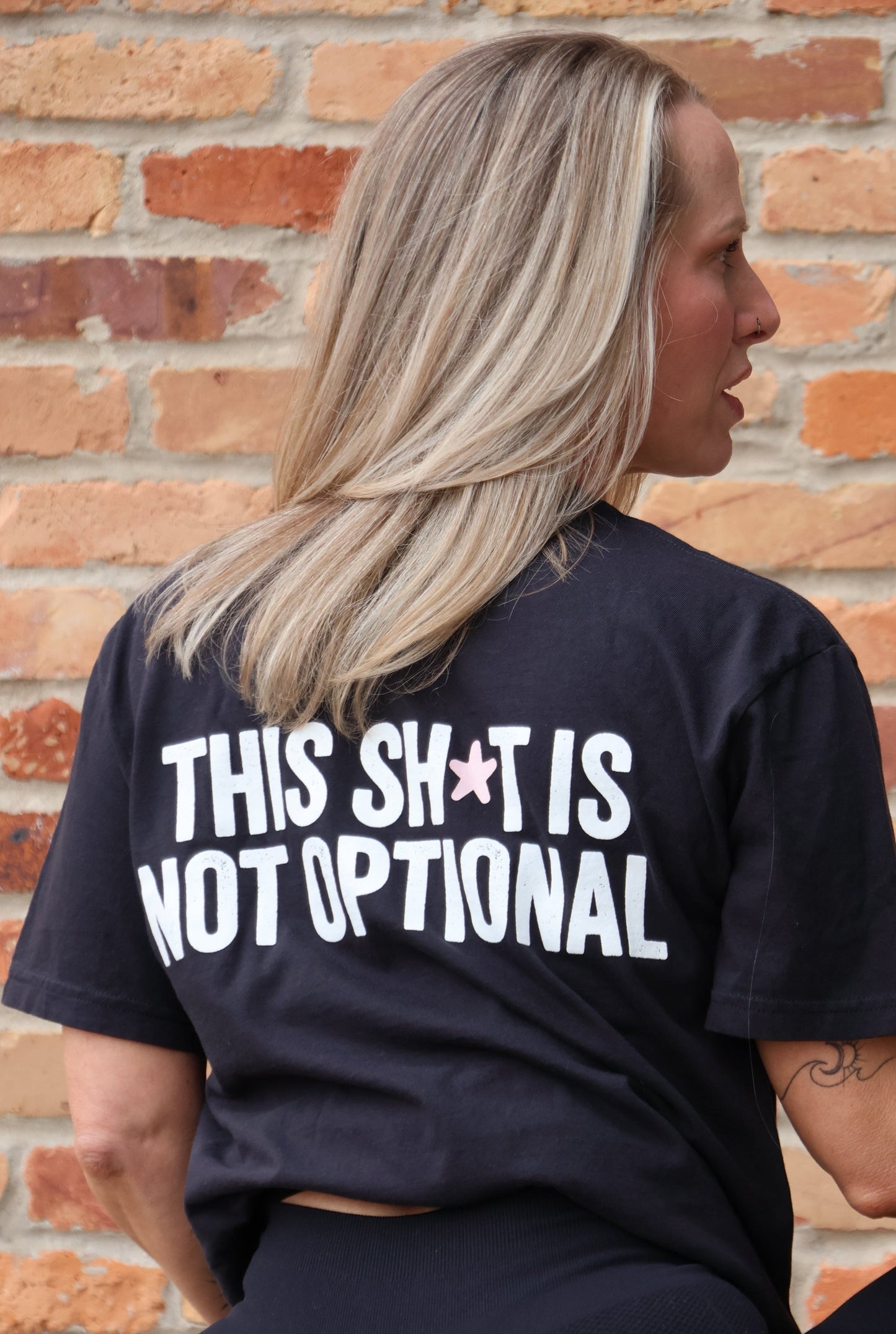 Person wearing a black t-shirt with 'THIS SH*T IS NOT OPTIONAL' text against a brick wall.